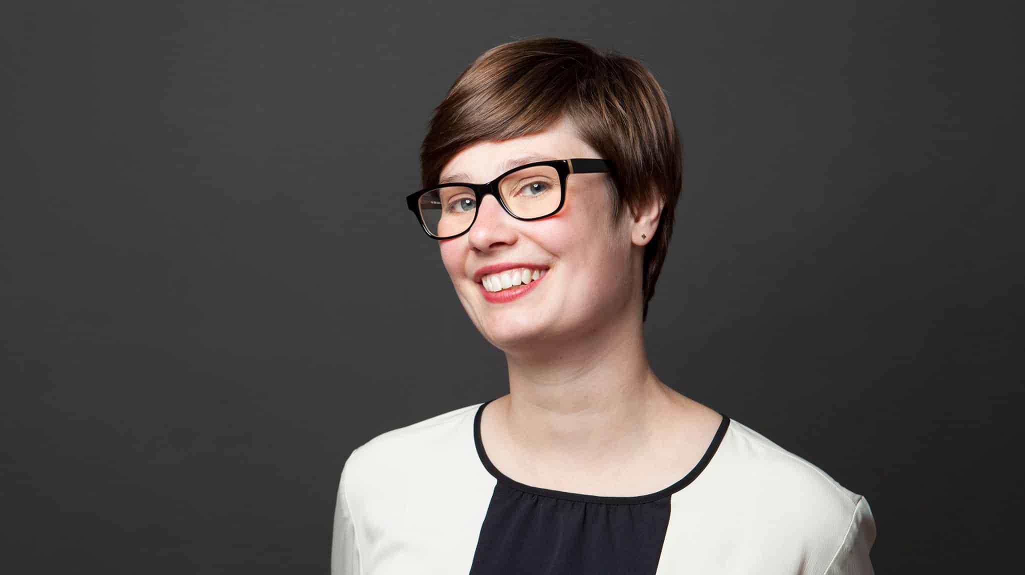 Die junge Gründerin Bianca Limbach hält ihre Kopf leicht schräg und lächelt offen. Sie trägt eine gerahmte Brille und trägt ihr braunes Haar kurz. Sie posiert vor einem dunklen Hintergrund und trägt ein helles Kostüm.