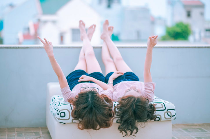 Zwei Frauen liegen auf einer Terrasse dem Rücken und heben sie Beine auf das Geländer. Beide sehen sich ähnlich, sie haben beide lockige braune Haare und tragen blaue Shorts. Sie strecken jeweils einen Arm in die Luft und blicken nach oben.