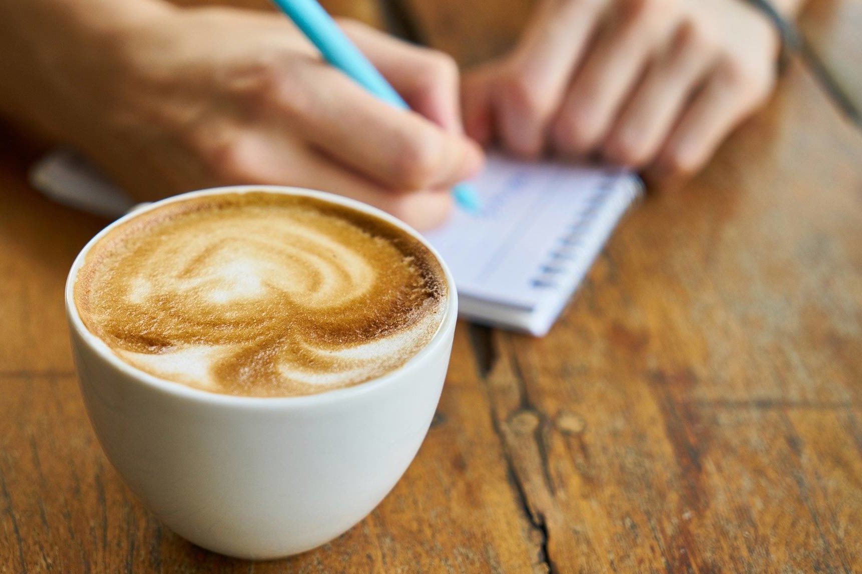 Eine Kaffeetasse mit Milchschaum. Dahinter eine Hand, die einen Stift hält und etwas in ein Notizheft schreibt.