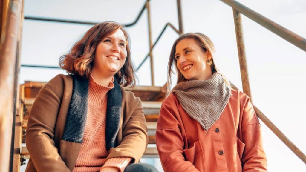 Zwei junge Frauen lächeln, die linke Frau schaut dabei schräg nach oben rechts, sie hat schulterlange, gewellte, braune Haare, trägt eine dicke braune Jacke und einen Pullover in orange, die rechte Frau lächelt die linke direkt an, sie hat lange, glatte, blonde Haare, trägt einen Mantel in orange und ein graues Tuch um den Hals