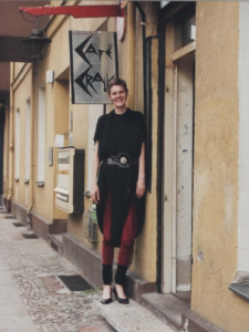 große, junge Person mit Pumphosenkleid, Leggings, Stulpen und Pumps steht lächelnd vor einem Eingang unter einem Schild, auf dem in spitzen, eckigen Buchstaben Café Cralle steht.