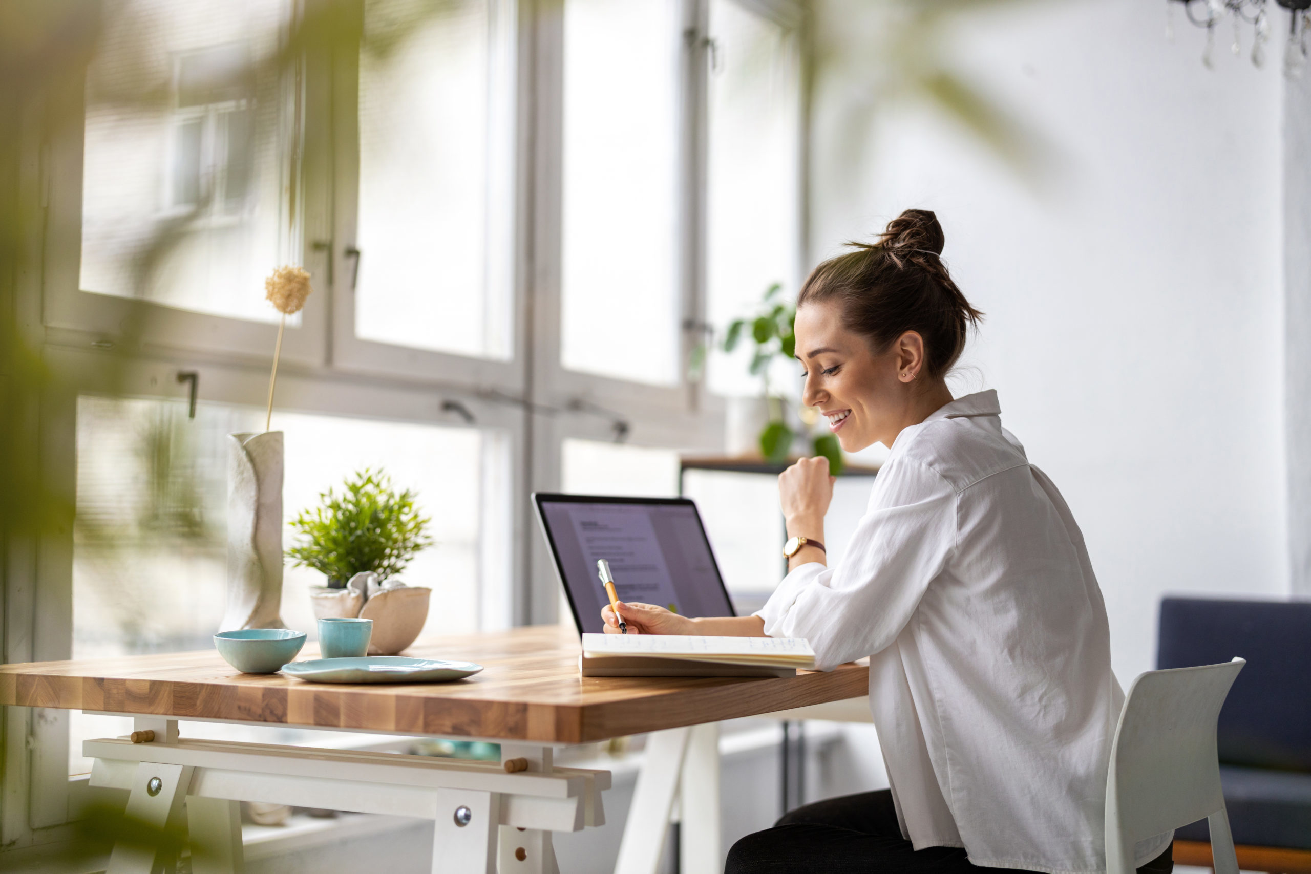 Frau mit weißer Bluse und Dutt sitzt vor Laptop am Schreibtisch, der an großer Fensterfront steht. Sie lächelt, hält einen Stift und schaut auf ihren Notizblock.