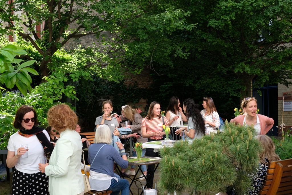 Frauen stehen verteilt und in Gruppen im Grünen und sind vertieft in Gesprächen. Sommerliche Atmosphäre. Tische stehen verteilt im Gelände.