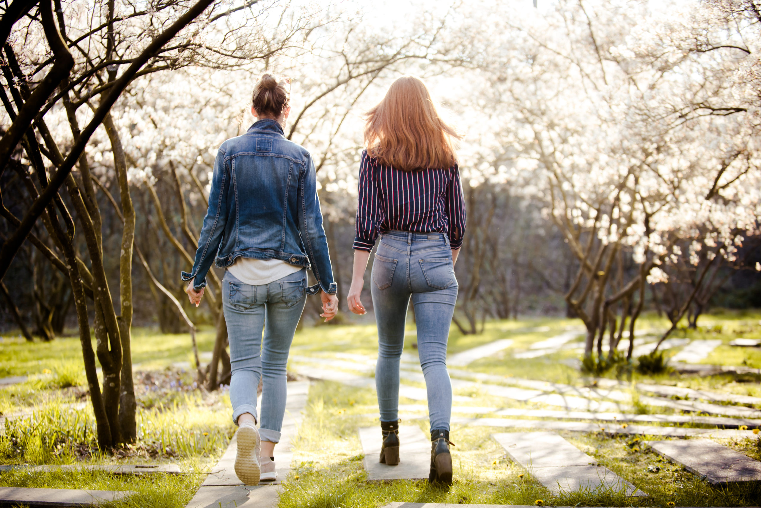 Naturumgebung mit Betonweg. Zwei Frauen spazieren gemeinsam, mit dem Rücken zur Kamera gerichtet. Beide tragen helle Jeans, die eine eine Jeansjacke, die andere eine leichte, dunkle Bluse.