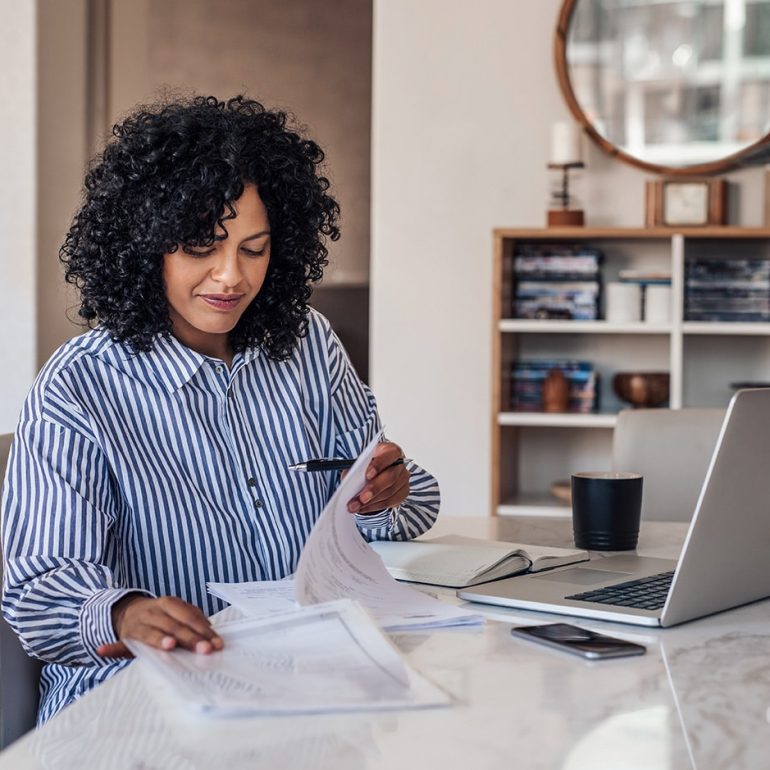 Smiling young female entrepreneur going through paperwork while working on a laptop at her dining room table at home