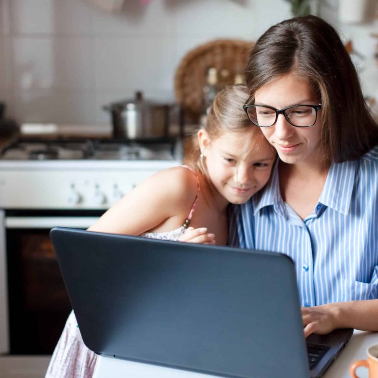 Mother and daughter using laptop and Internet. Freelancer workplace in cozy kitchen. Woman and child girl together. Concept of female business, working mom, freelance, home office. Lifestyle moment.