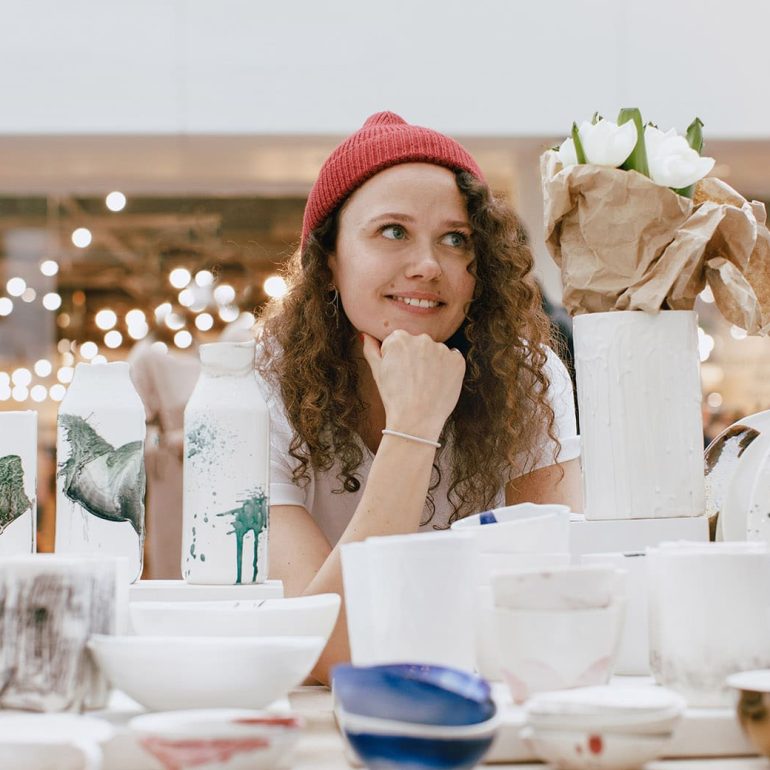 Young hipster woman selling behind counter with her ceramics and porcelain hand-painted at local market of craftsmen, small business